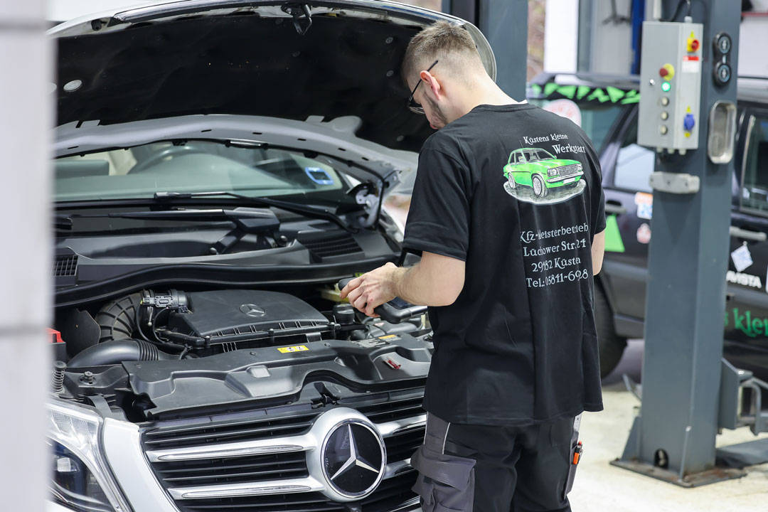 Ein KFZ-Mechatroniker von Küstens kleine Werkstatt in Küsten führt mit einem Diagnosegerät eine Fehlersuche im Motorraum eines Mercedes-Benz Transporters durch. Er trägt ein T-Shirt mit dem Firmenlogo.